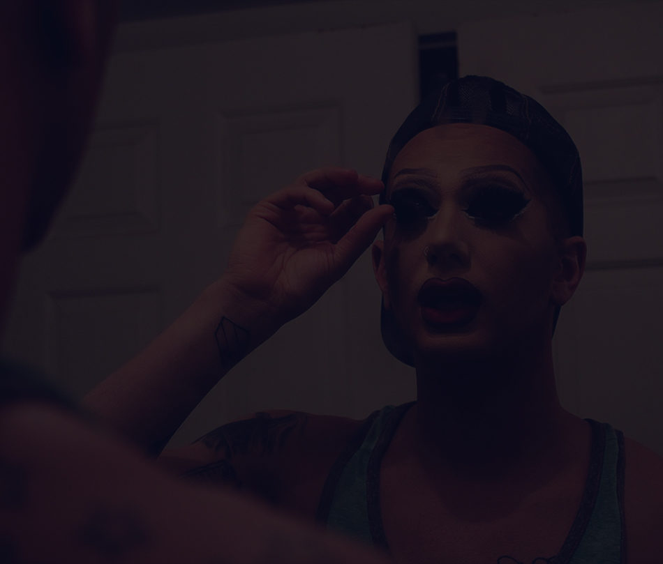 A photo of Matty Cameron, applying makeup in the mirror. There is a dark overlay over the photo.