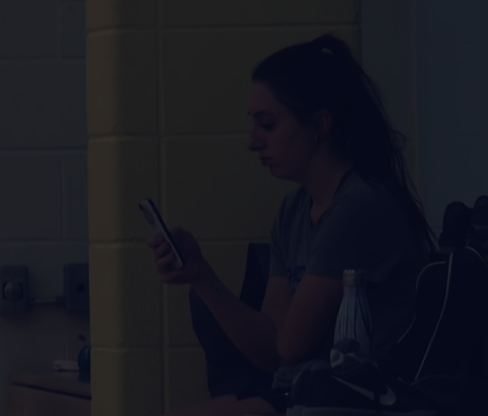 A person is sitting, looking at their phone, with a yellow wall in the background. There is a dark overlay over the photo.