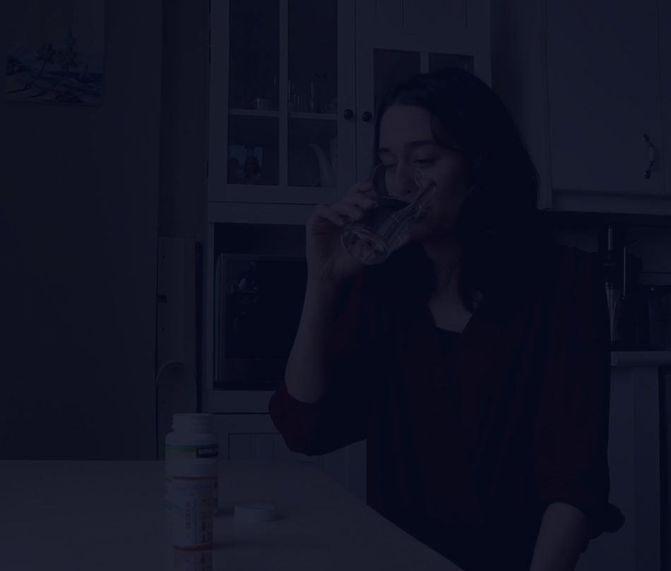 A person is standing in a kitchen drinking a glass of water, with a container of something open on the counter. There is a dark overlay over the image.