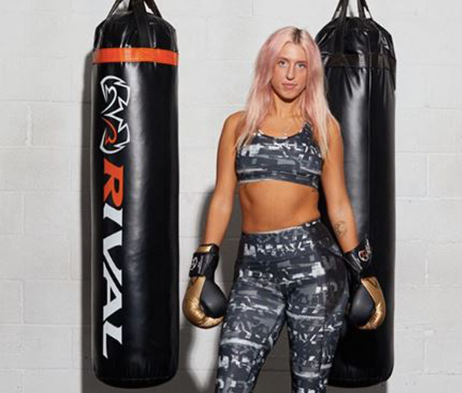Someone wearing athletic gear and boxing gloves, standing in front of two punching bags and a white bacgkround.